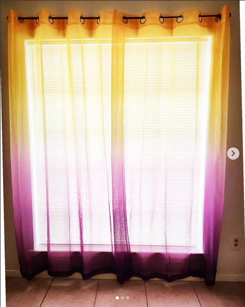 Image of curtains hung in a window. The curtains are sheer and dip dyed, yellow at the top half and purple at the bottom half.